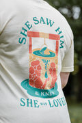 Beige t-shirt with a graphic design and text on the back, worn by a person.